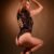 Side view of a woman in a black lace bodysuit and stiletto heels, arms raised above her head against a warm brown backdrop.