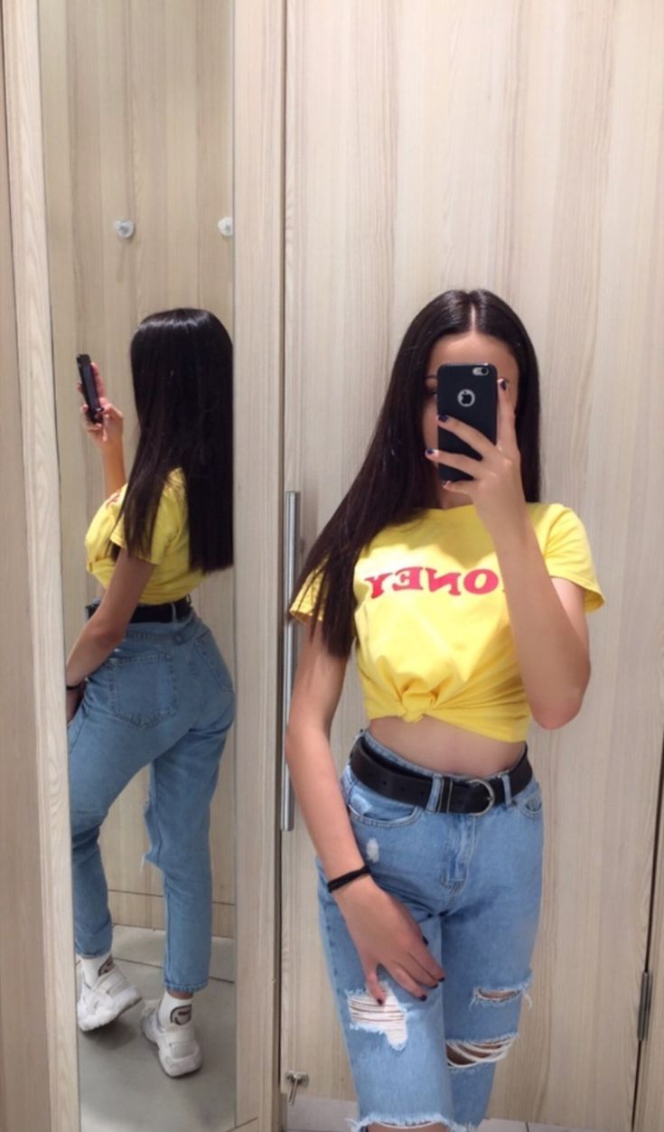 Woman takes a mirror selfie in a fitting room, wearing a yellow cropped tee and ripped jeans with a black belt.