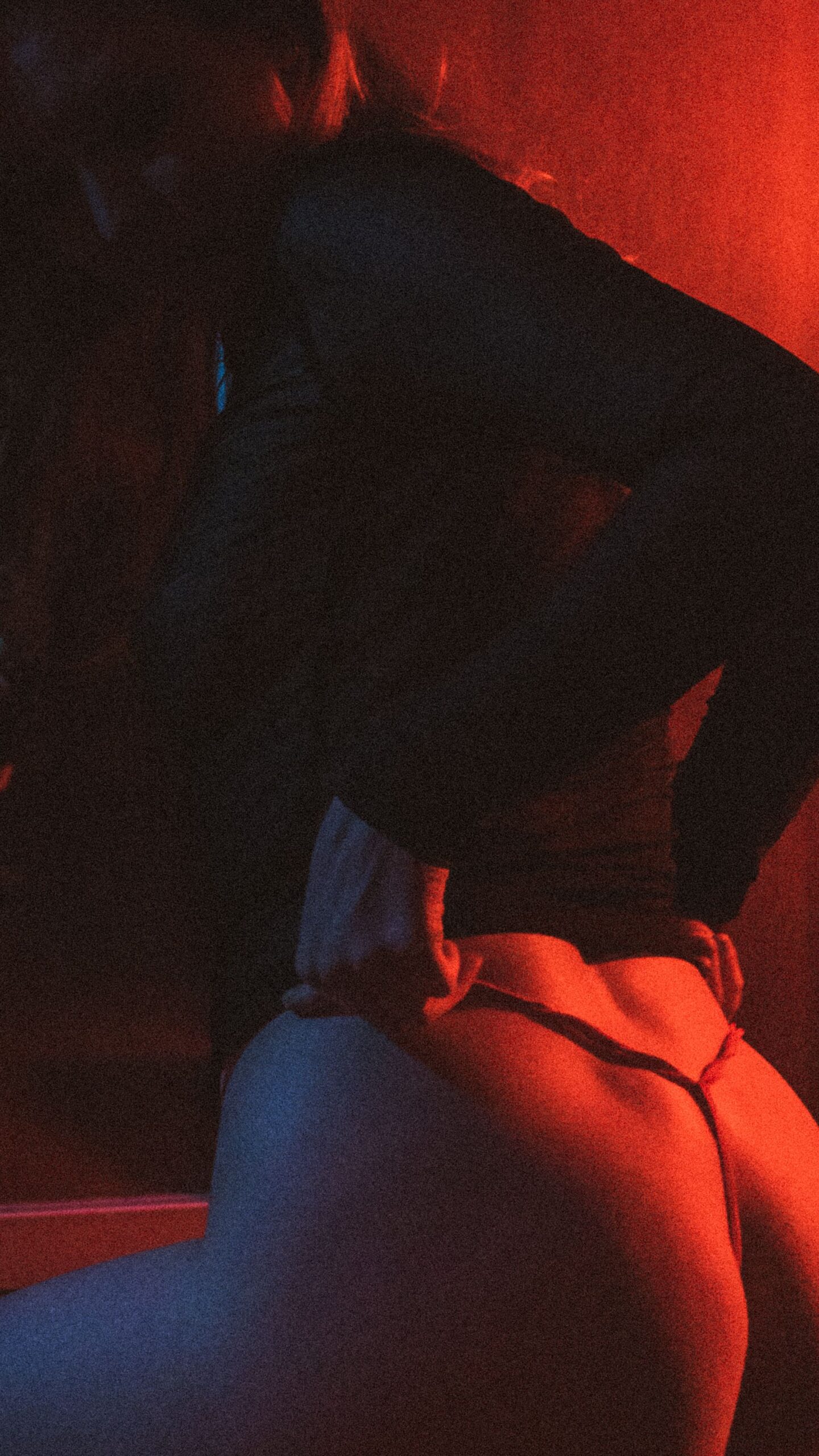 Person wearing a hoodie seen from behind in a dim red-lit setting, appearing to hug someone nearby.